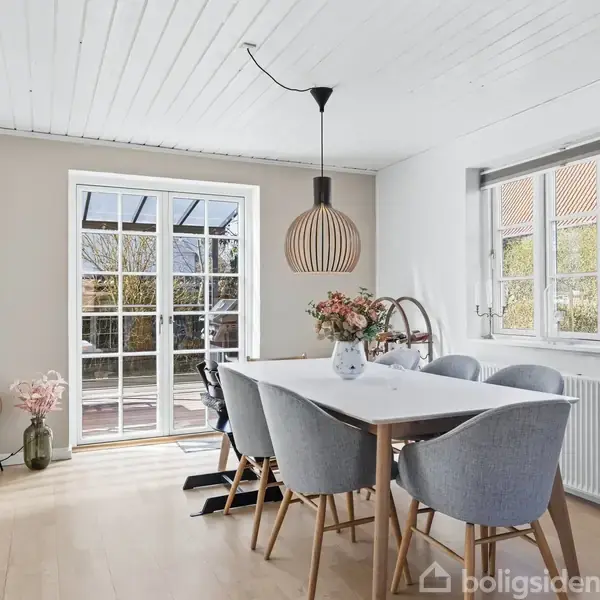 Et spisebord med seks stole står i et lyst rum. En lampe hænger over bordet, og der er blomster i en vase. Store vinduer giver udsigt til en terrasse.