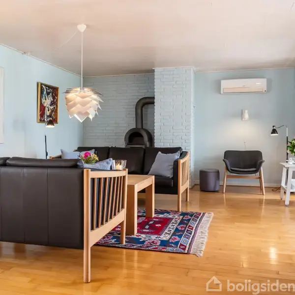 Sort sofasæt i en lys stue med trælaminatgulv, farverig tæppe, pejs, og kunstværker på væggene. Der er store vinduer, et lille bord med blomster og en moderne loftslampe.