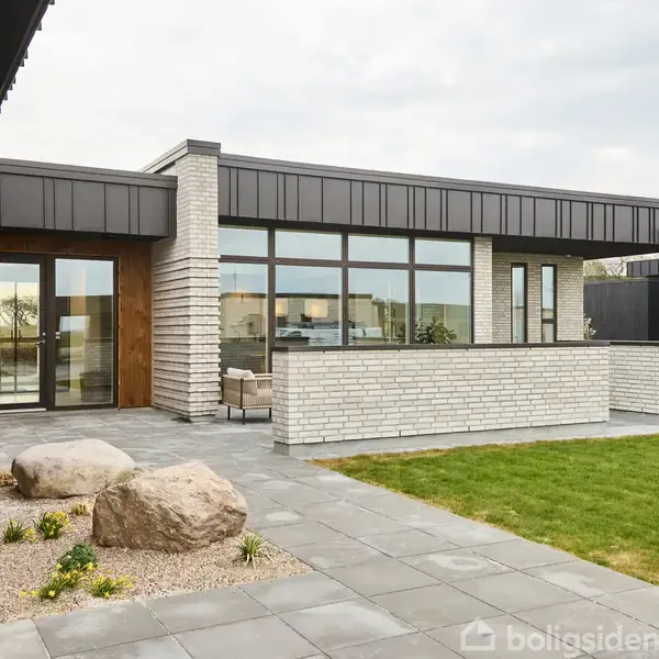 En moderne bygning med store glasvinduer står som fokus. Den er omgivet af en flisebelagt terrasse, sten og græsplæne, hvilket skaber en stilren og åben atmosfære.