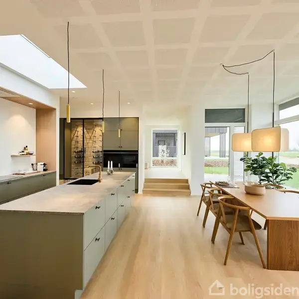 Et moderne køkken med en stor ø-arbejdsbordssektion, omgivet af grå skabe. Til højre er der et spisebord med stole og en plante ved siden af store vinduer, der giver udsigt til en have.