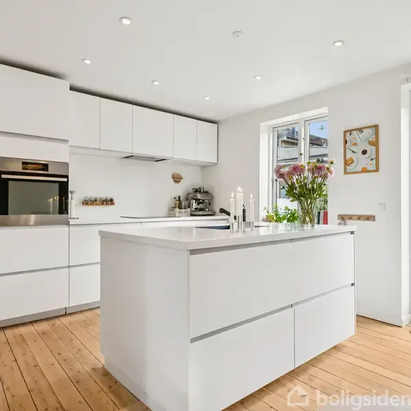 Et moderne køkken med hvide skabe og en central ø. Bordpladen har lysestager og friske blomster. Trægulve og store vinduer giver rummet naturligt lys. En have ses udenfor.