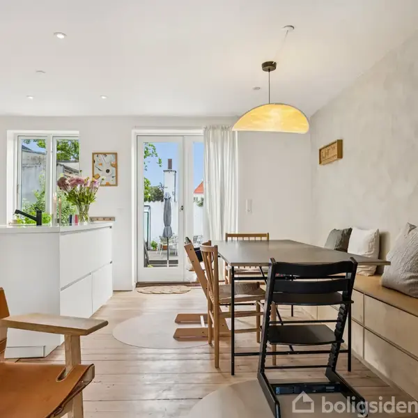 Spisebord med stole står i en lys, moderne spisestue. Bænken ved siden af bordet har puder, og rummet har store vinduer, der giver udsigt til en solrig terrasse.