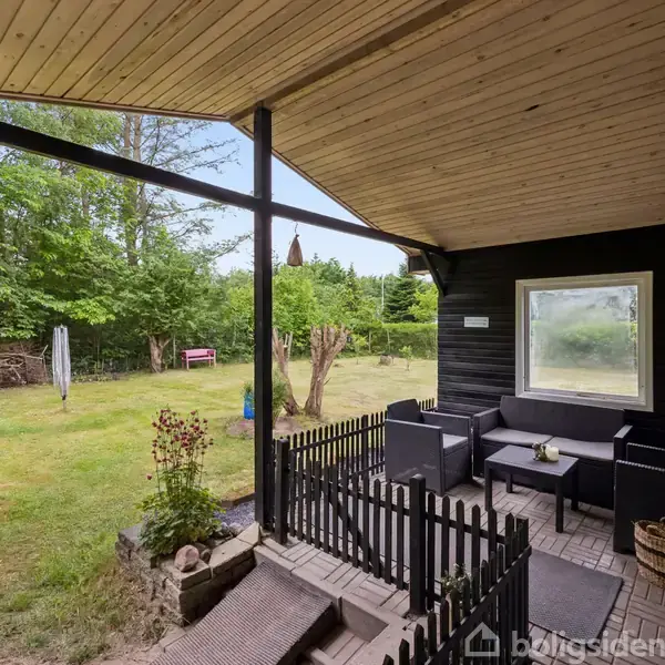 Terrasse med sortmalede møbler ved et bord; omgivet af træhegn. Græsplæne og buske ses i baggrunden. Træterrasse overdækket med naturligt træloft, med udsigt til grøn have.
