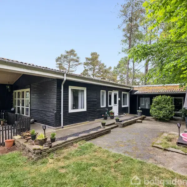 Sort træhus står stille i en frodig have med brostensbelagt terrasse. En lyserød børnepool står på terrassen ved siden af et bord med parasol omgivet af høje træer.