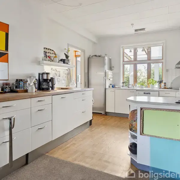 Et moderne køkken med hvide skabe udstyret med kaffemaskine og køkkenredskaber, placeret ved siden af et stort køleskab. Væggen prydes af farverige abstrakte malerier.