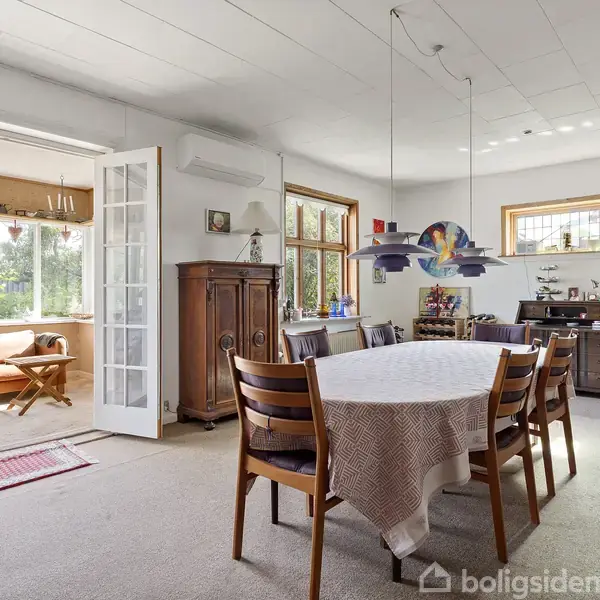 Spisebord med fire stole dækket af dug står i en rummelig og lys spisestue. Bagved åbner franske døre ud til en veranda med sofa og udsigt til grøn have.