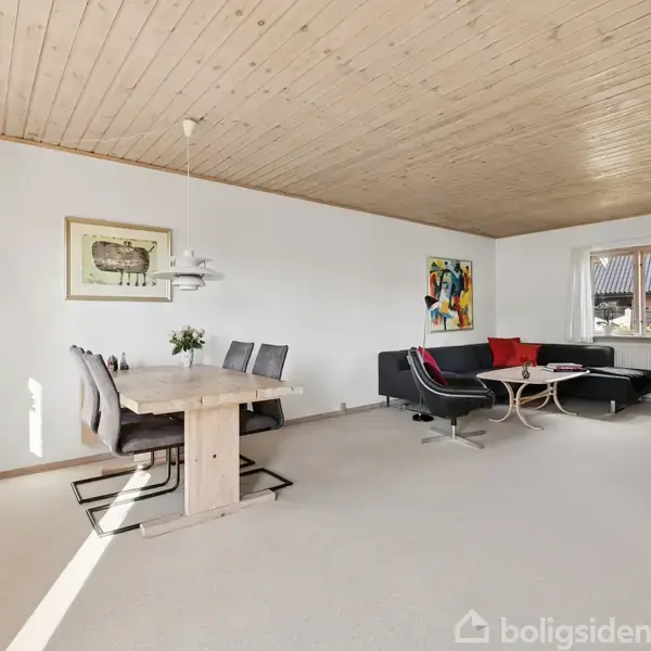 Spisestue med lyse trægulve og loft, en spisebord med fire stole, sofaområde med sort sofa og røde puder, bogreol, malerier på væggene. Meget naturligt lysindfald.