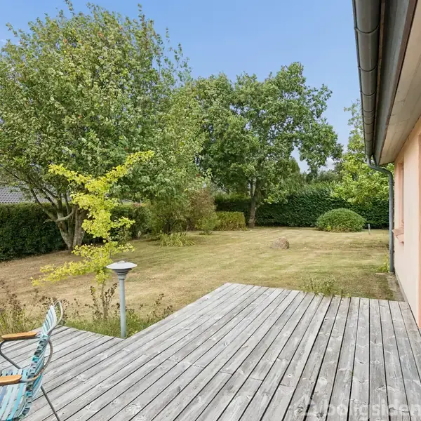 Træterrasse med runde havebord og stole. Omgivet af en velplejet have med grønne buske og træer. Huset har store vinduer, og græsplænen er ved siden af terrassen.