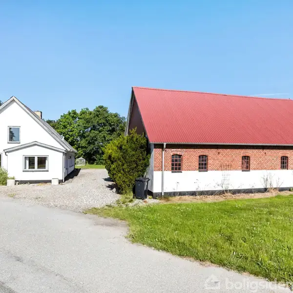 Hus til venstre, hvidt med mørkt tag, står stille. Rød murstenslade med rødt tag til højre. Omgivet af græs og træer under blå himmel.