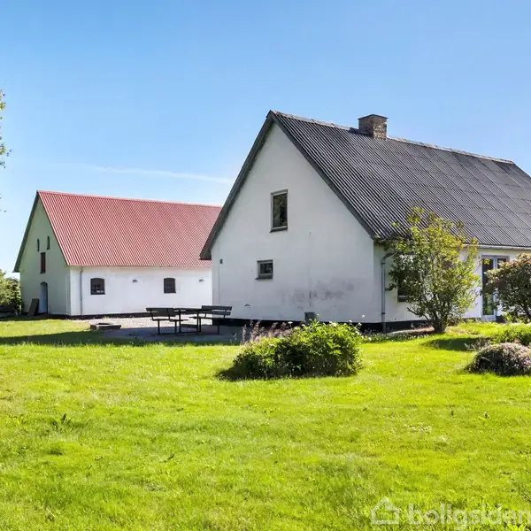Et hvidt hus med gråt tag står i en grøn have med en større bygning med rødt tag i baggrunden. Det er omgivet af træer og buske under en klar blå himmel.