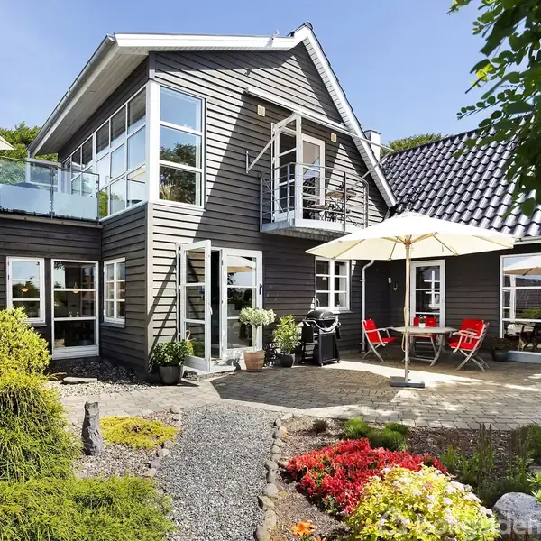 Et moderne, sort-hvidt to-etagers hus med fladt tag omgiver en terrasse med parasol og røde stole. Omkringliggende have har stier, buske og farverige blomster.