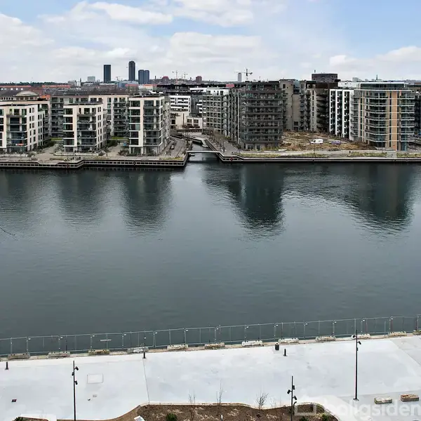 Flod flyder roligt mellem moderne boligbygninger i et byområde, hvor en promenade løber langs vandet. Det fjerne bybillede ses i baggrunden under en let overskyet himmel.