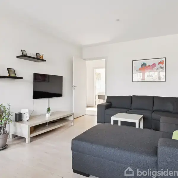 En stor, mørkegrå sofa står i en stue med hvide vægge. Et TV hænger på væggen over en lav reol, og der er en plante i en potte ved siden af. En dør står åben til et andet rum. En farverig plakat hænger på væggen.