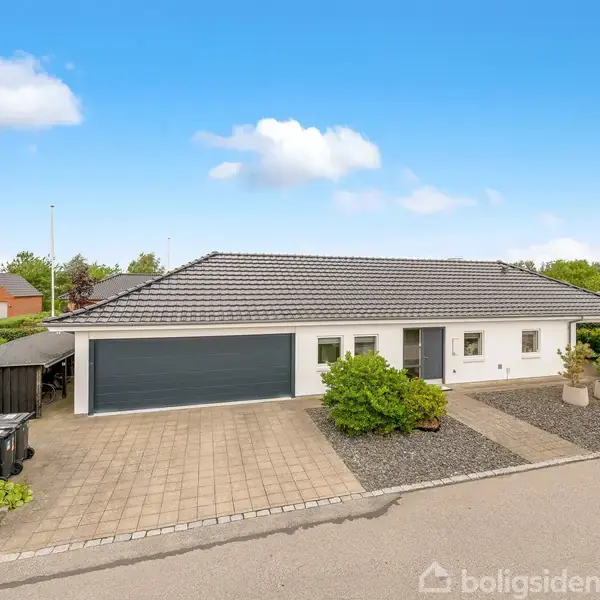 En moderne etplans villa med gråt tag og hvid facade står på en brolagt indkørsel. Den har en dobbeltgarage, omkranset af grønne buske og grus, i et boligkvarter.