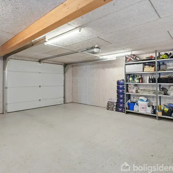 En rummelig garage med en åben, tom gulvplads. På højre side står reoler fyldt med forskellige værktøjer og genstande til opbevaring. Loftet har lysstofrør.