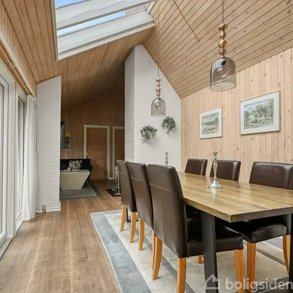 Langbord omgivet af seks mørke stole står på et tæppe i en spisestue med højt til loftet. Rummet har trægulve, trævægge, store vinduer og skrålofter med ovenlys.