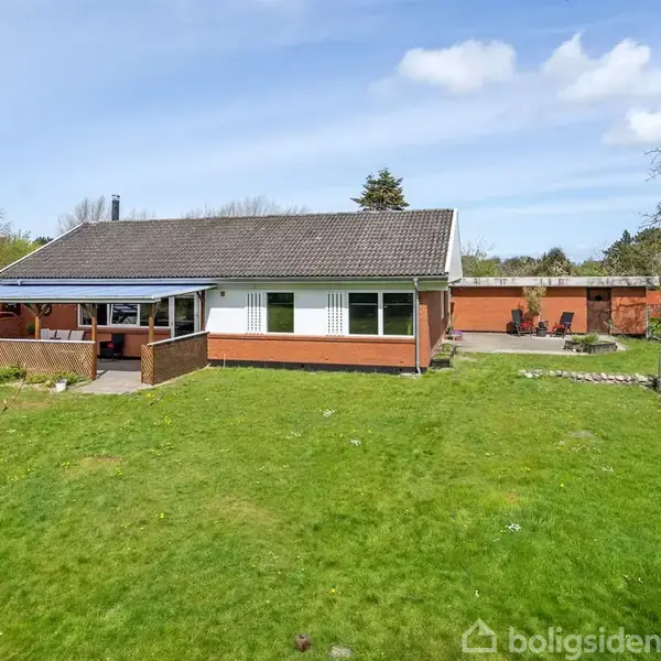 Et enkelt hus med hvid facade og sort tag står på en stor græsplæne. Haven er omkranset af træer og en sti fører til en terrasse.