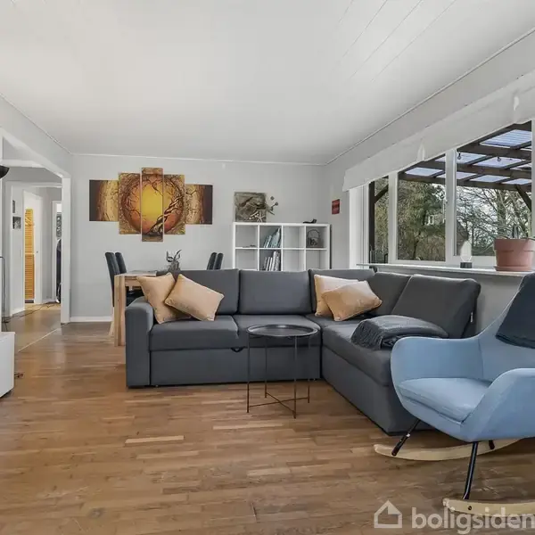 En stue med en grå hjørnesofa og blå lænestol ved et sofabord. Væggene er hvide, og rummet har store vinduer med udsigt til en have. Et TV står på en medieenhed.