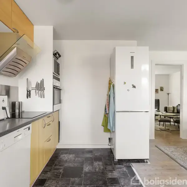 Køkken har lys træindretning, opvaskemaskine og køleskab; i baggrunden ses en stue med sofa og stol. Gulvet er sort og hvidt flisemønster. Dørene er hvide.