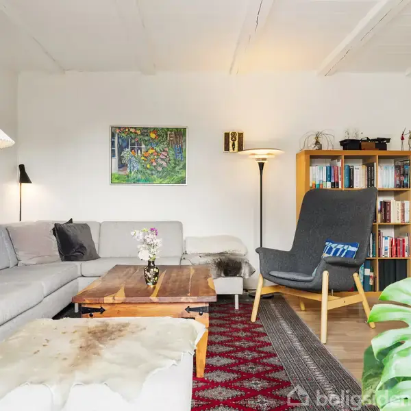 Grå sofa står i en stue ved et stort træbord med blomster. En lænestol er ved siden af en bogreol fyldt med bøger. Grønne planter er i forgrunden.