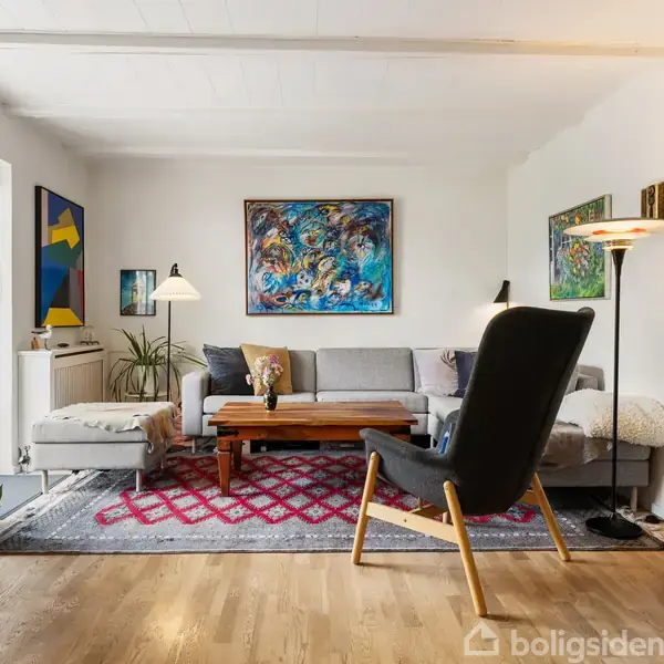 En stue med en grå sofa og stol på et rødt mønstret tæppe. En bogreol og kunstmalerier pynter væggene. Bordlamper og planter skaber en hyggelig atmosfære.