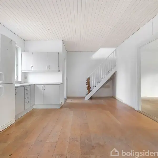 Køkken i et moderne hus med lyse træskabe og stålkomfur; en åben trappe fører opad; rummet har lyse vægge og trægulv.