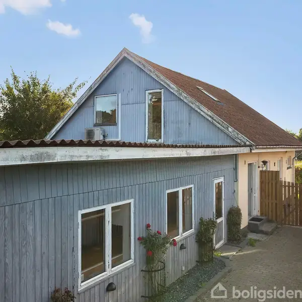 Træhus med blå facade og rødt tag står i solskin. Foran huset er der en lille gårdsplads med grønne planter. Huset har flere vinduer og en indgangsdør.