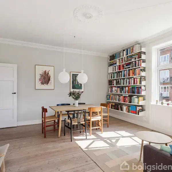 Spisebord med stole står midt i et lyst rum med hvide vægge og trægulv. Bogreol fyldt med bøger ved siden af stort vindue. To hængelamper over bordet.