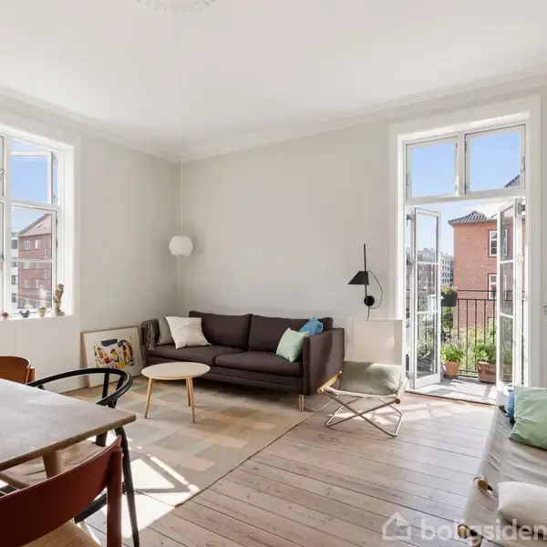 Stue med en brun sofa i hjørnet, sofabord og lænestol ved siden af. Store vinduer og altandør lukker lys ind. Trægulv og et spisebord med stole i forgrunden.