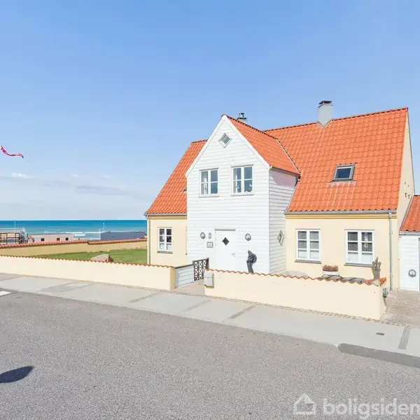 Et gult hus med rødt tegltag står stille i et kystområde ved havet. En dansk flagstang står ved siden af huset, og en vej løber foran.