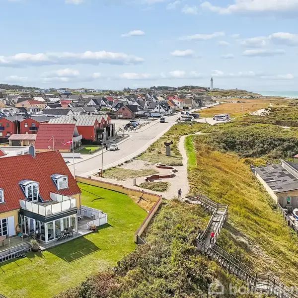 Hus med rødt tag står nær kysten, med velholdt have foran. Kystlinje og hav strækker sig i baggrunden, omgivet af flere huse og grønne områder under en blå himmel.