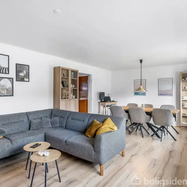 En grå hjørnesofa med gule puder står ved et sofabord i en lys stue. Billeder hænger på væggen; reoler tilfører opbevaringsplads. I baggrunden står et spisebord med stole.