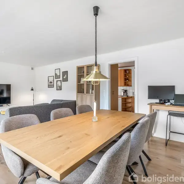 Spisebord med seks grå stole står i en lys stue. En lampe hænger over bordet. I baggrunden er der en sofa, fladskærms-TV, skrivebord og computer.