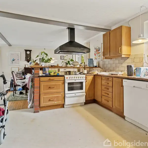 Et køkken med træskabe og opvaskemaskine. En ovn med emhætte står centralt, omgivet af diverse køkkenredskaber. Til venstre ses en spiseplads nær et stort vindue.