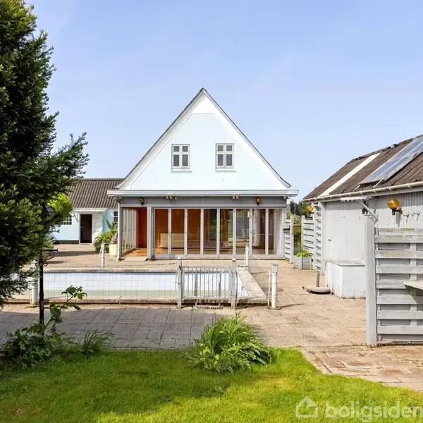 Hvidt hus med trekantet tag, terrasse og store vinduer, omkranset af have og flisebelagt område. To sidelænger med træfacader i nærheden. Ingen synlig tekst udover Boligsiden vandmærke.