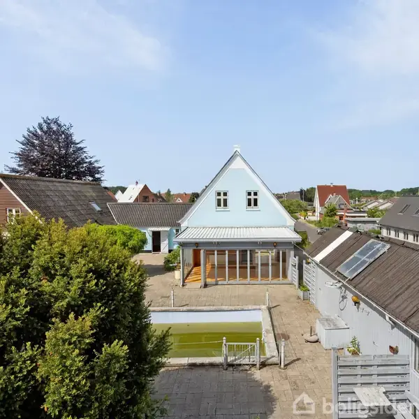 Et lille, gråt hus med blågrønt tag står for enden af en gårdhave med en pool. Omgivet af andre huse og en frodig have under en klar himmel.