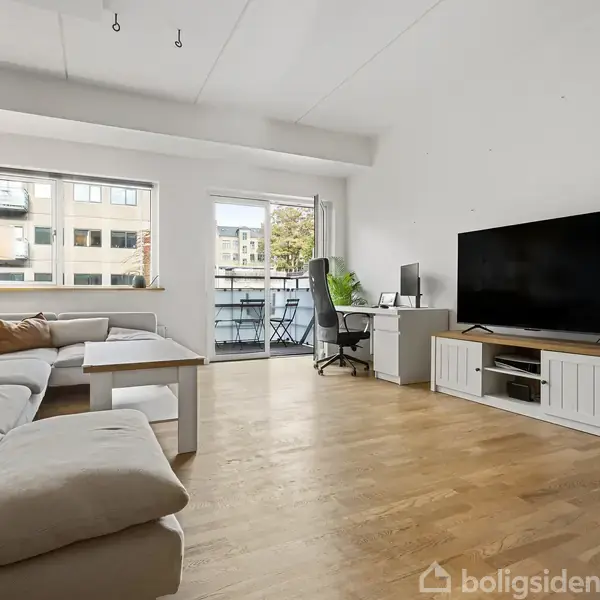 En stue med lys sofa arrangeret rundt om et sofabord. Action: Tv'et står på et skab ved siden af en arbejdsstation. Context: Store vinduer giver udsigt til bygninger udenfor.