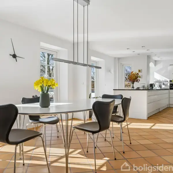 Rundt spisebord med sorte stole i moderne, lyst køkken. På bordet står en vase med gule blomster. Rummet har terracotta-gulv og store vinduer, hvilket skaber et lyst miljø.
