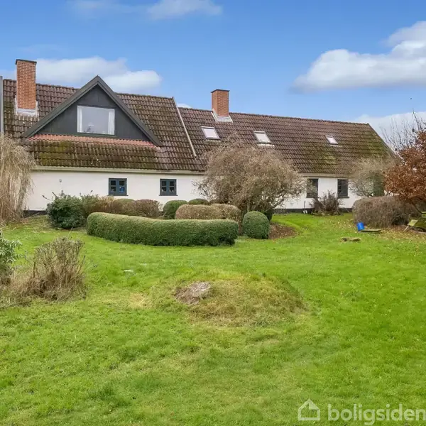 Et stort hus med skråtag og kvistvindu i midten står omkranset af velplejet græsplæne og buske. Skyfri himmel og træer ses i baggrunden.