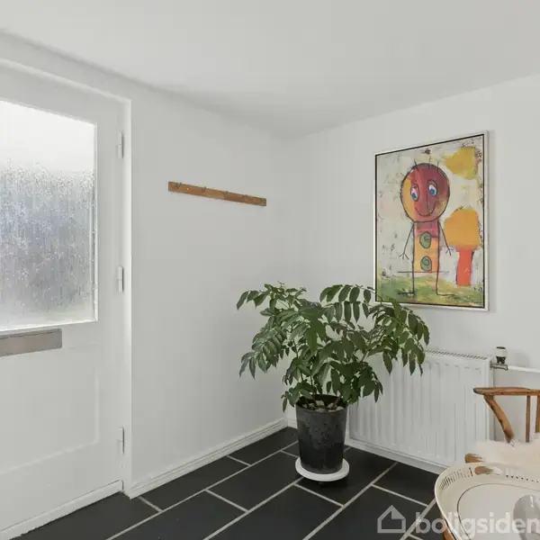 Dør med matteret glas står stille i en lys entre. Ved siden af er en grøn plante og en stol. En farverig plakat hænger på væggen.