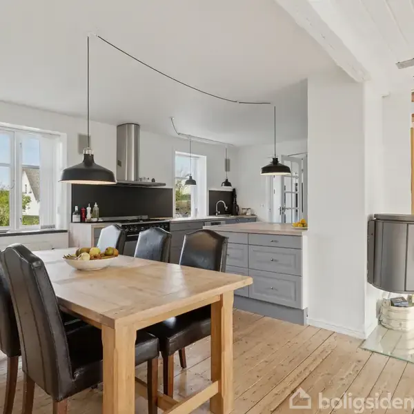 Et lyst køkken med træbord, omgivet af sorte stole, to lamper hængende over bordet. Køkkenet har grå skabe og en ovn. Store vinduer giver udsigt til træer udenfor.