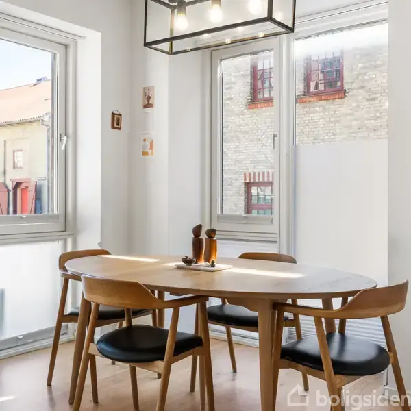 Ovalt spisebord med fire stole, placeret i et lyst hjørne med store vinduer. Moderne loftslampe hænger over bordet. Væggene er dekoreret med små billeder. Ingen tekst fundet.