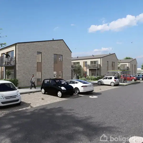 Flere moderne rækkehuse med fladt tag, bygget af mursten, står langs en vej. Biler er parkeret foran husene, og en person går på fortovet. Græs og træer i baggrunden.
