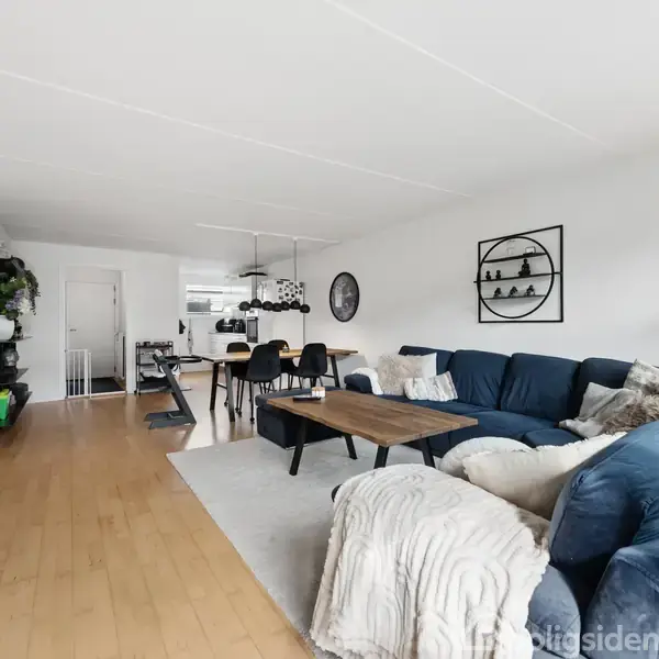 En stor stue med en blå hjørnesofa fyldt med puder og et tæppe. Et træbord står foran sofaen. Rummet har lyse vægge og trægulve, med en spiseplads i baggrunden.