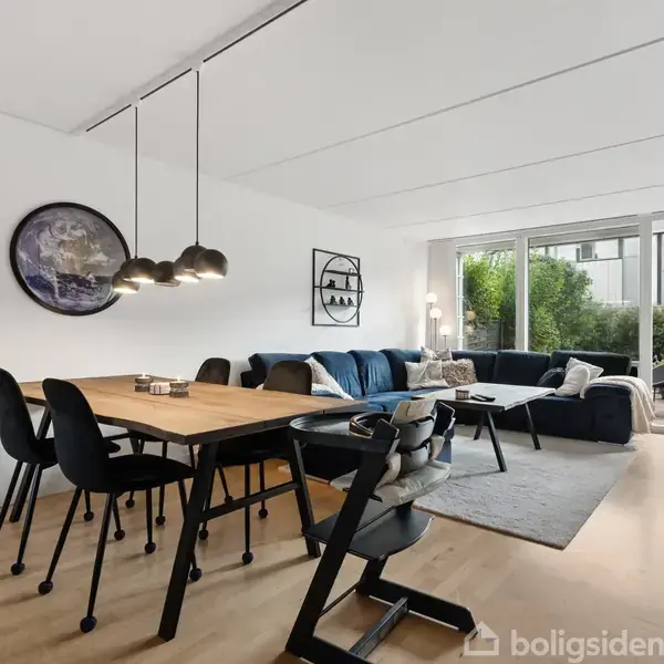 Spisebord med sorte stole står i en stue med trægulv. Stuen har en stor, blå sofa, et tæppe, og store vinduer, der åbner ud til en terrasse med udsigt til haven.