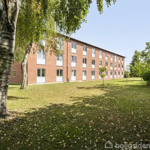 En rød murstensbygning står med flere vinduer, omgivet af en græsplæne med træer i forgrunden, der skaber en skyggefuld atmosfære.