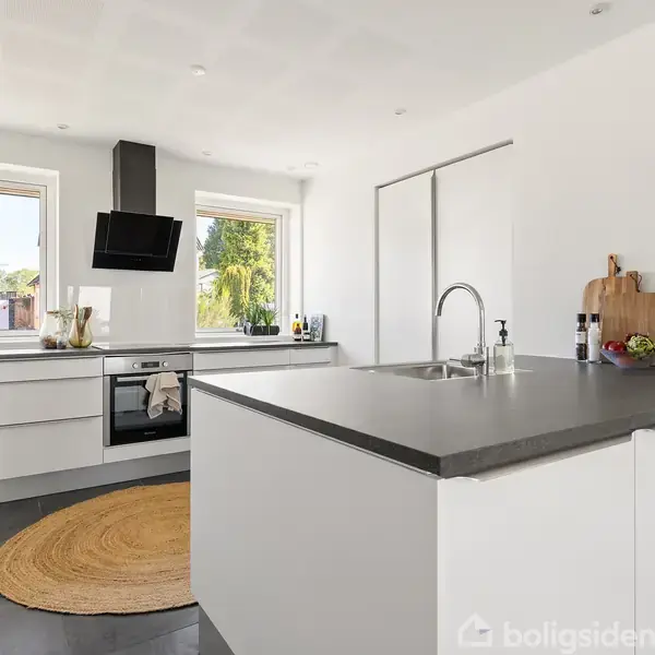 Moderne køkken med hvidt køkkenbord og sort bordplade, central vask og træskærebræt. Lyst interiør med to vinduer, sorte fliser, rund tæppe, indbygget ovn og ventilator.