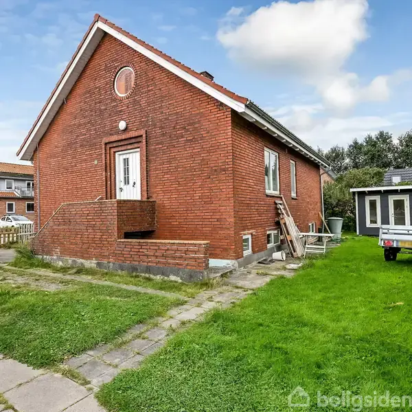 Rødt murstenshus står stille i en grøn have med en hvid dør og vinduer. Den omkringliggende have har en trailer og et skur, beliggende i et boligområde.