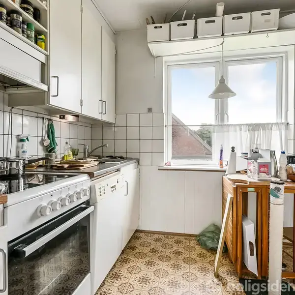 Et køkken med hvide skabe og en komfur med ovn. Kogegrej og krydderier er synlige. Et lille bord står ved et vindue med udsigt.