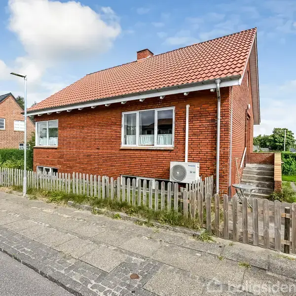 Rødt murstenshus med hvidt tag, placeret på en stille villavej. Foran huset er et lavt hegn og en luftvarmepumpe. Til højre er en indgang med en lille trappe.